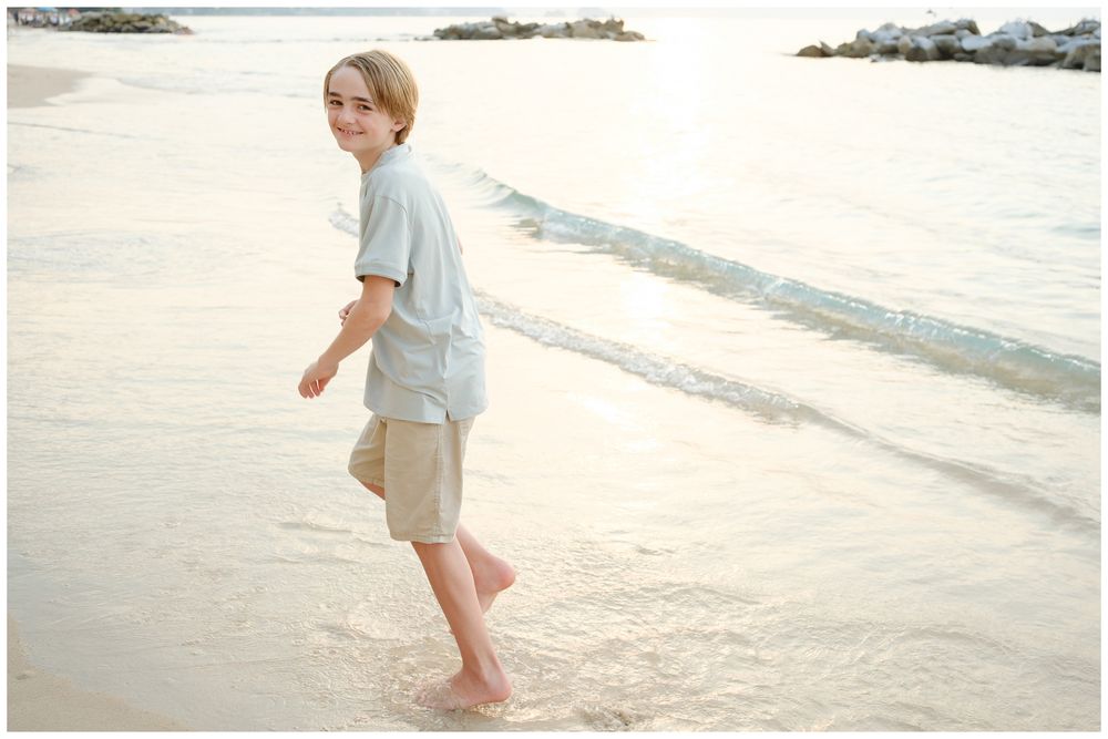 A smiling boy walks barefoot along a sandy beach shore, wearing a light blue shirt and khaki shorts.