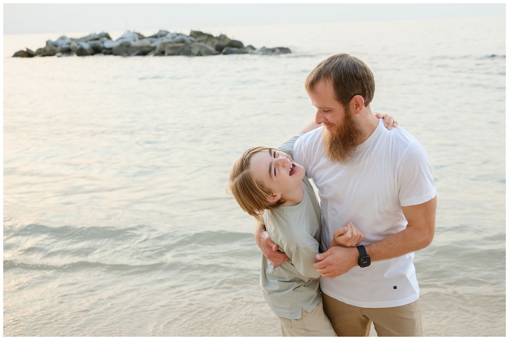 Couple laughing and embracing on a beach at sunset, man with beard hugging woman near rocky shoreline.