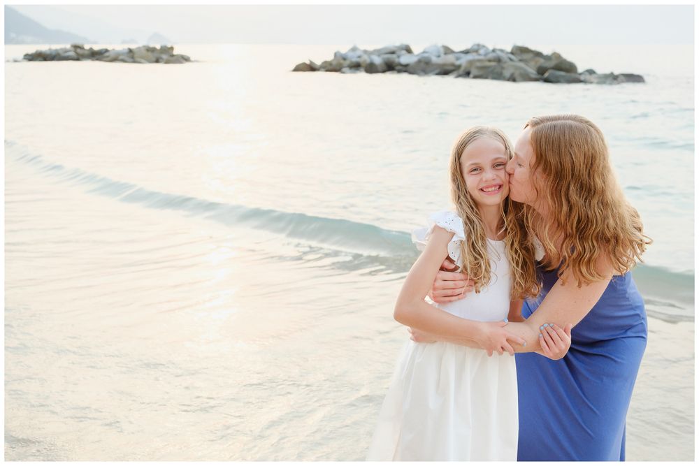 Two girls hugging and smiling on a sandy beach at sunset, one in white dress and one in blue.