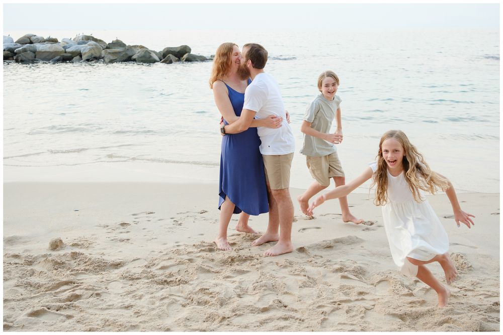 Family of four enjoying a beach day, parents kissing while children play in the sand by the ocean.
