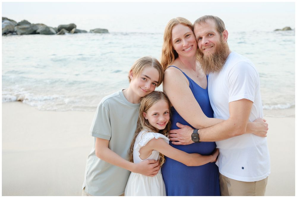 Happy family of four sharing a warm embrace on a scenic beach with rocky shoreline.