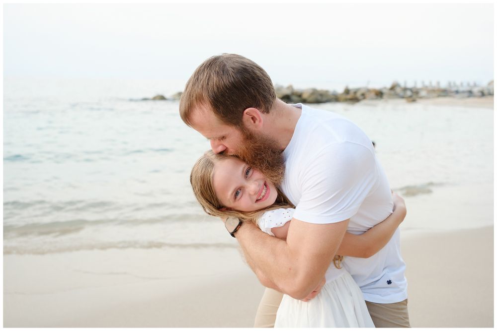 A couple shares a joyful embrace on a sandy beach, the man kissing the smiling woman's head near calm ocean waters.
