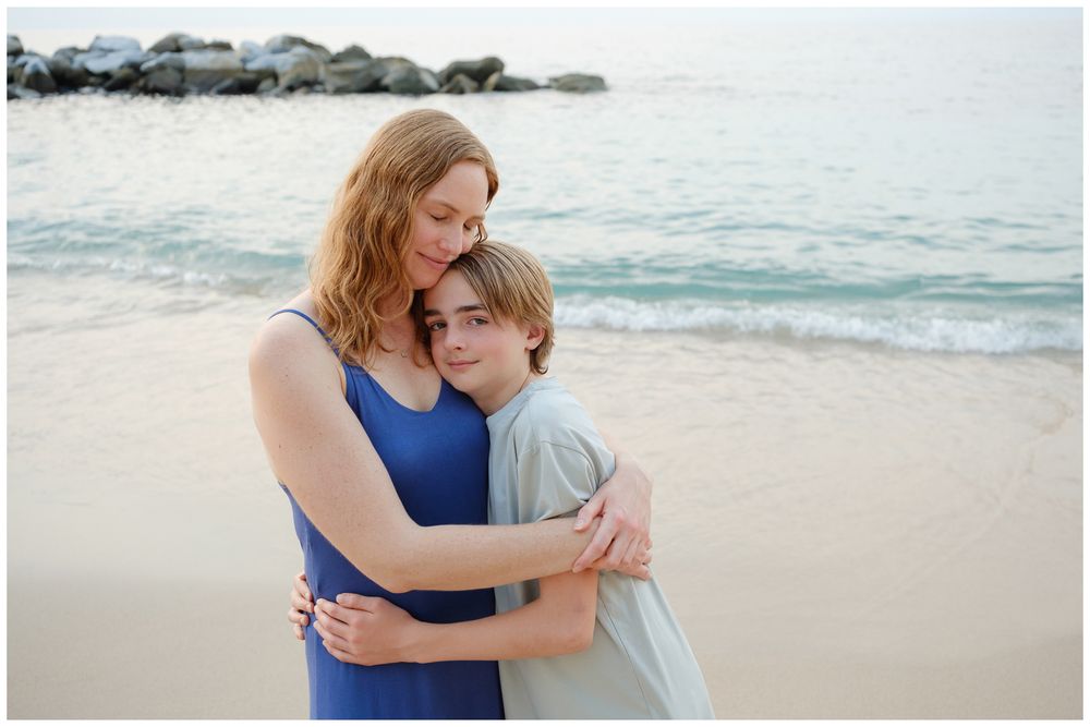 A mother and son share a warm embrace on a sandy beach with calm ocean waves in the background.