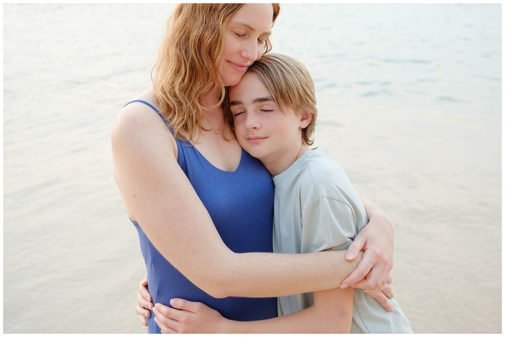A mother and young boy share a tender embrace on a beach, with calm water in the background.