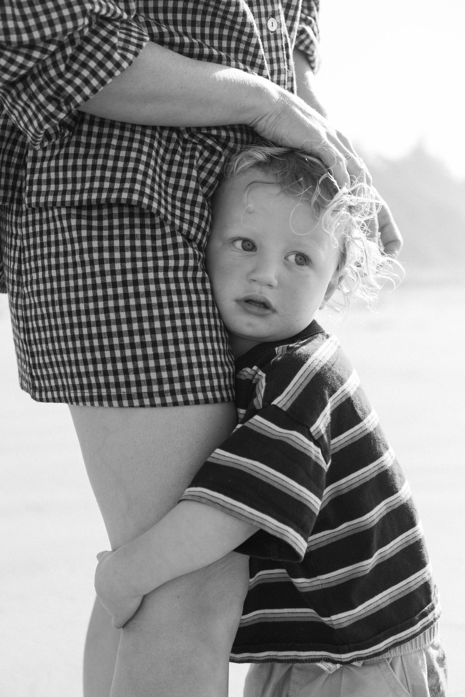 Black and white photo of curly-haired toddler clinging to parents leg outdoors.