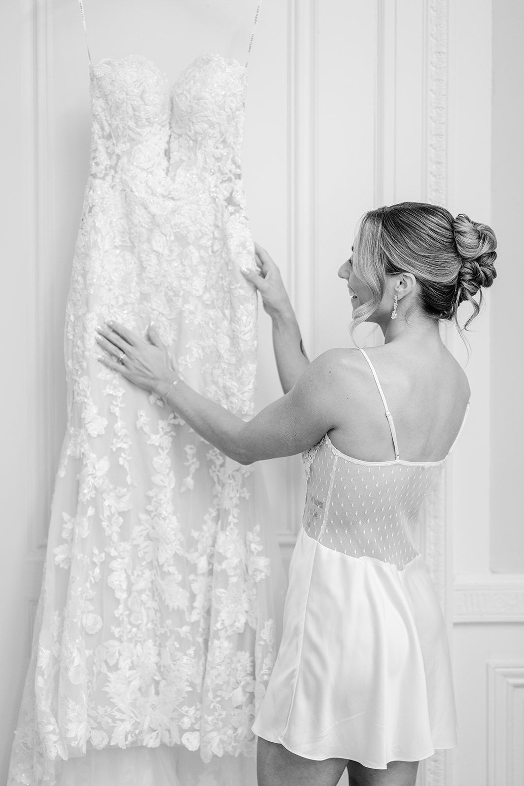 Armour House Wedding - Smiling bride in white slip dress admiring her floral lace wedding gown hanging in a bright room.