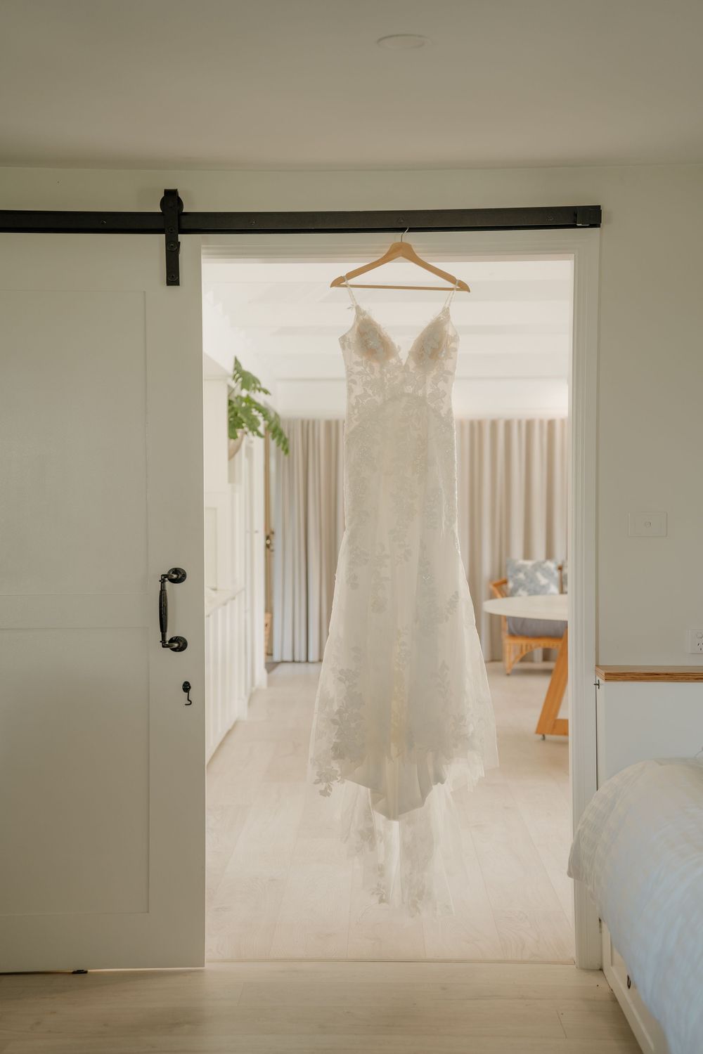Lace wedding dress hanging on wooden hanger in doorway of bright modern room with sliding barn door.