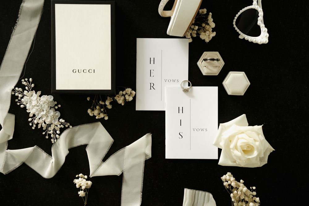 Elegant wedding flat lay featuring Gucci box, vow books, rings, pearl jewelry, white rose, and ribbon on black background.