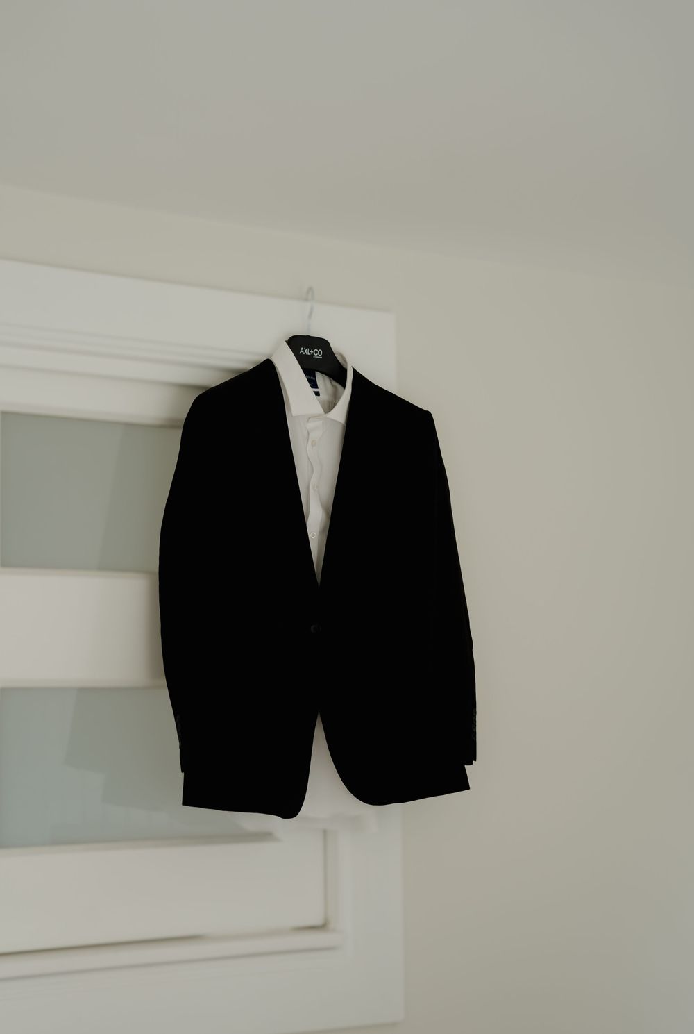 Black tuxedo jacket and white dress shirt hanging on door hanger in bright white room.