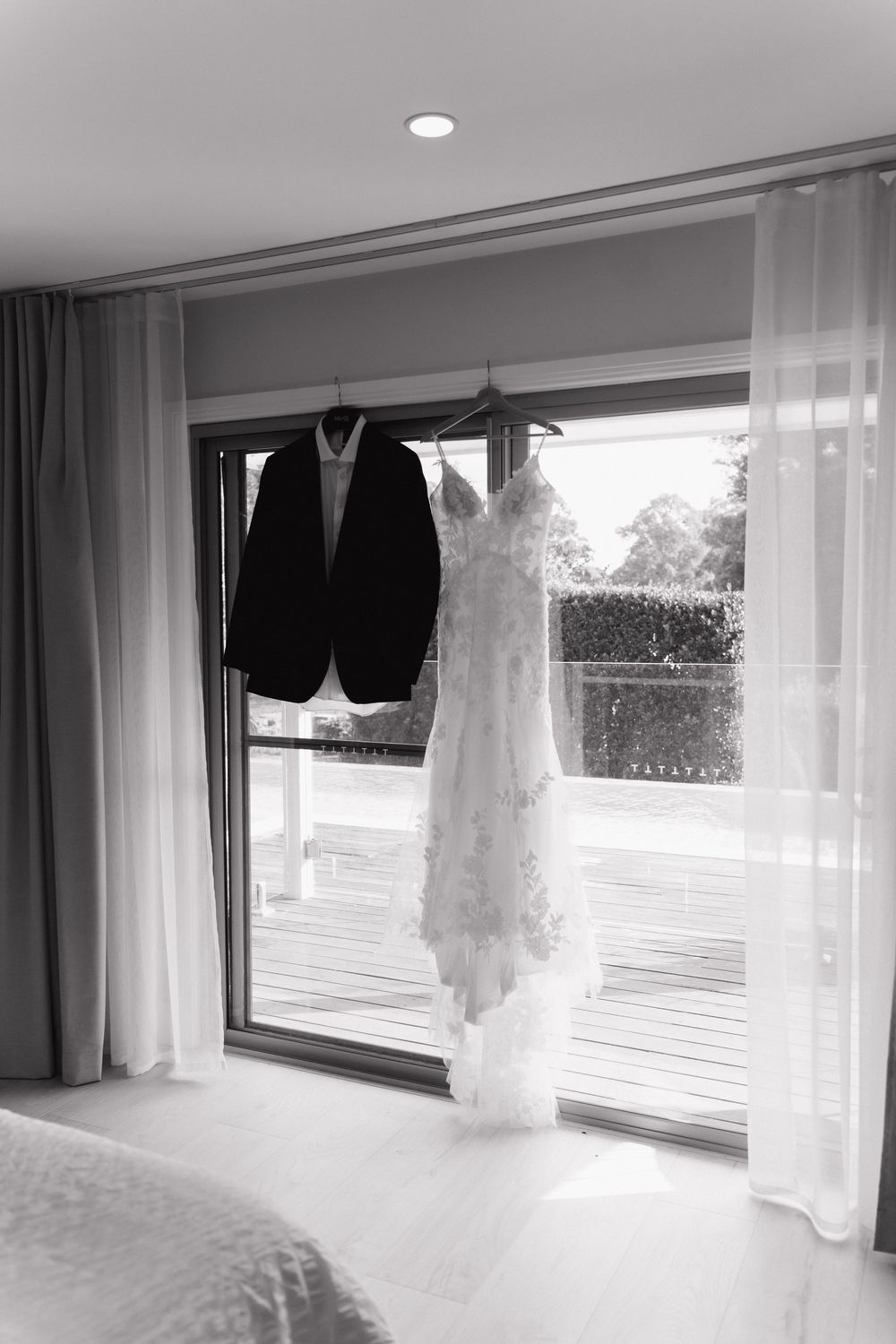 Black and white photo of a wedding dress and groom's suit jacket hanging together in a sunlit window.