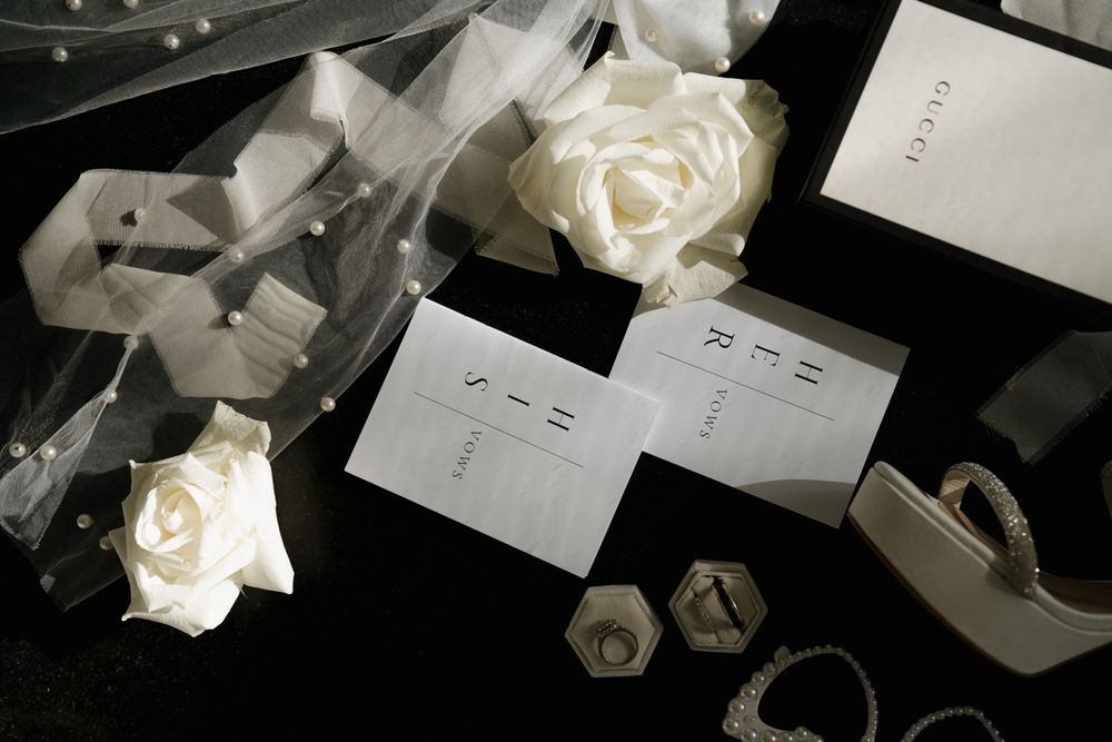 Elegant wedding flat lay featuring white roses, invitation cards, a veil, and jewelry on a dark background.
