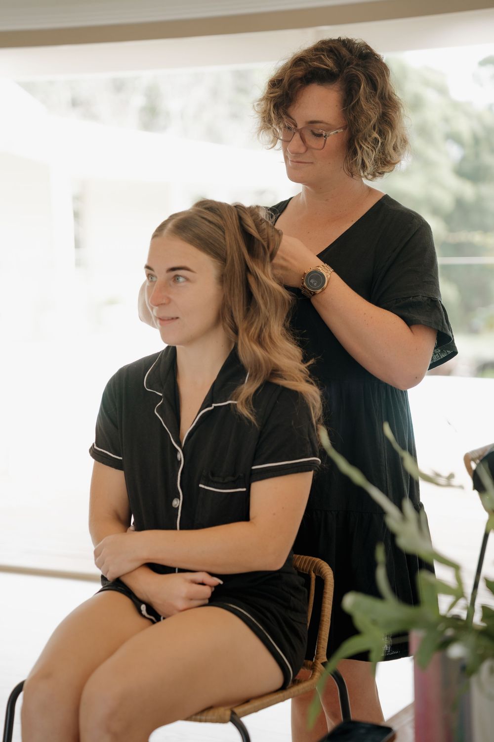 Hairstylist styling a young woman's long wavy hair in a black pajama set during bridal preparations.