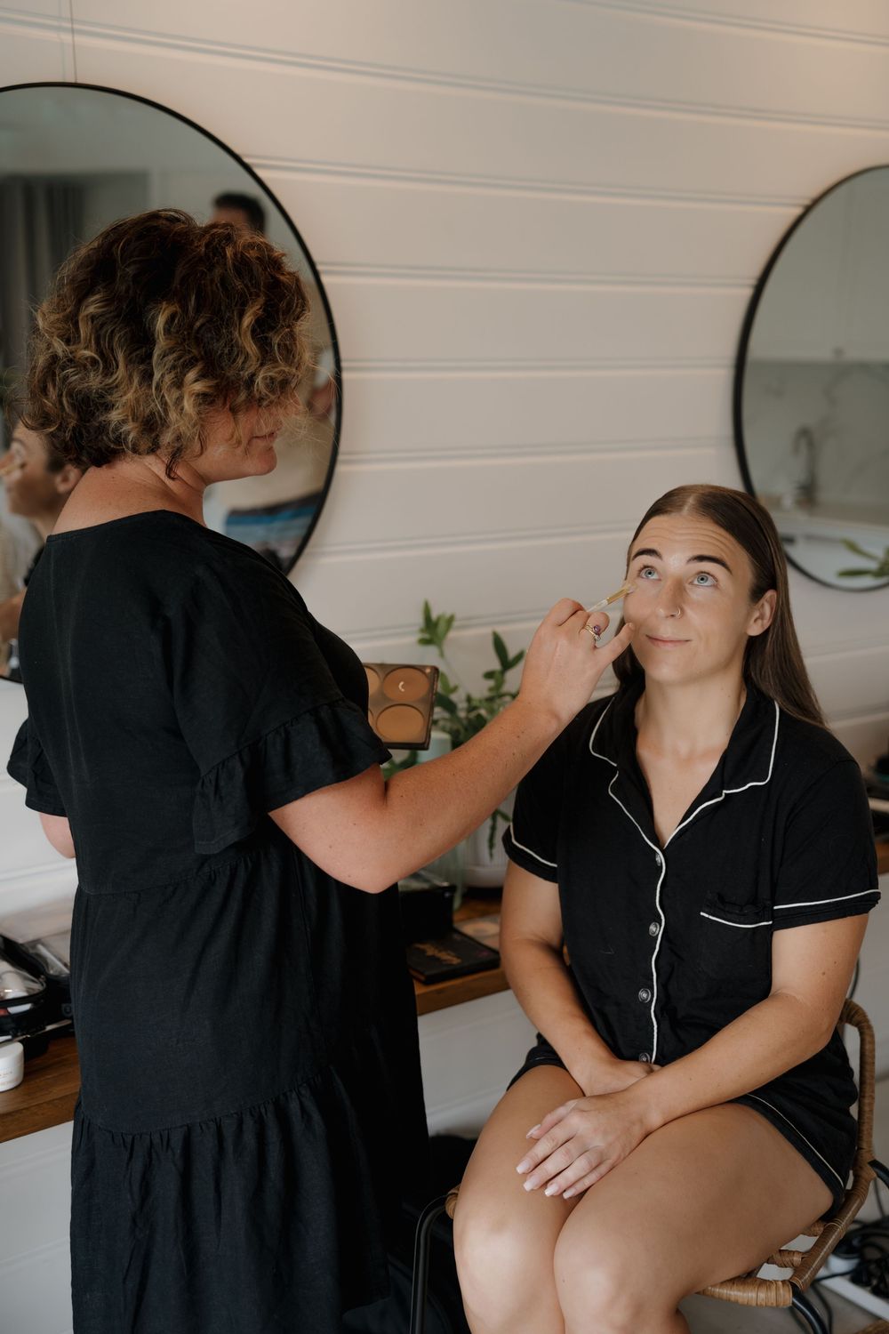 Makeup artist in black dress applying foundation to bride during wedding morning preparations.