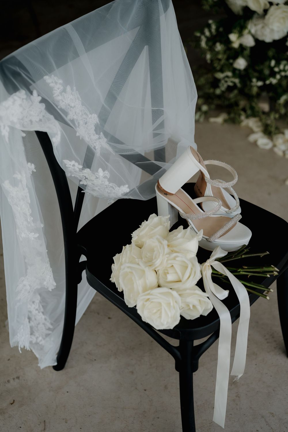 Bridal wedding details with white rose bouquet, strappy heels, and lace veil draped over a black chair.