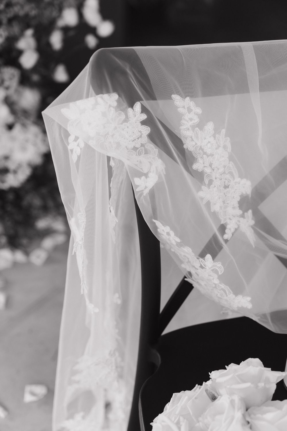 Black and white close-up of a delicate lace-trimmed bridal veil draped over a chair at a wedding ceremony.