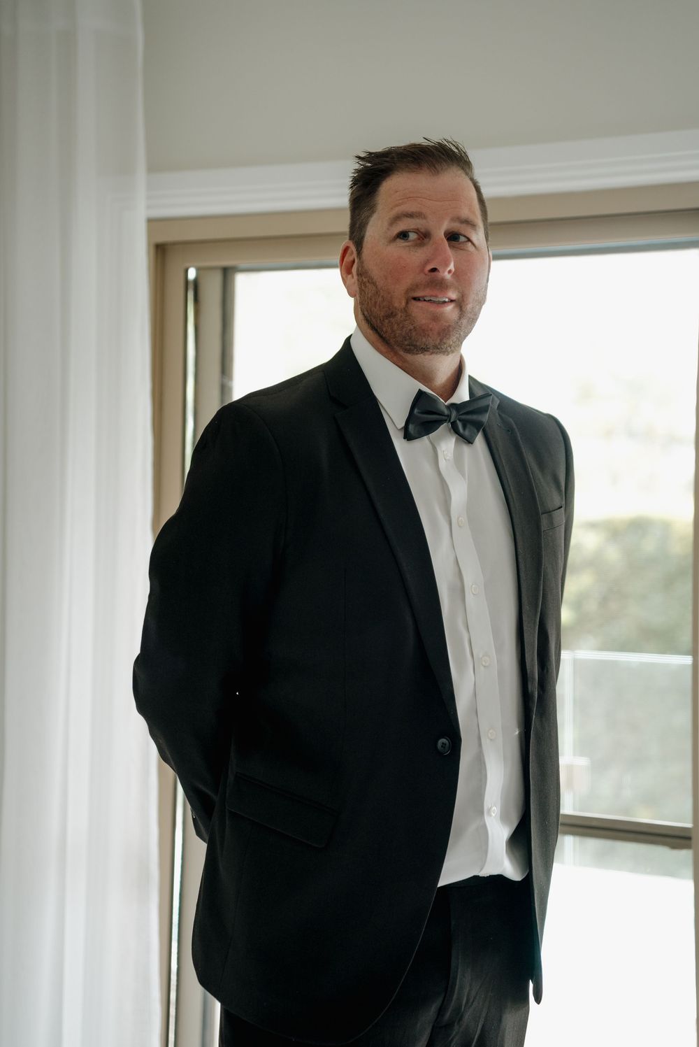 Handsome groom in black tuxedo with bow tie standing confidently by a bright window on his wedding day.