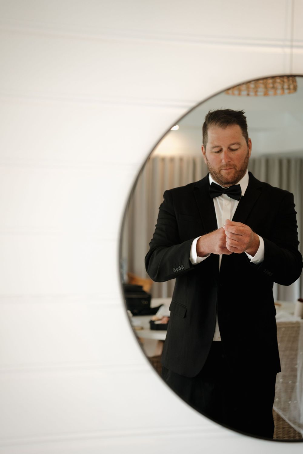 Groom in black tuxedo and bow tie adjusting cufflinks reflected in round mirror.