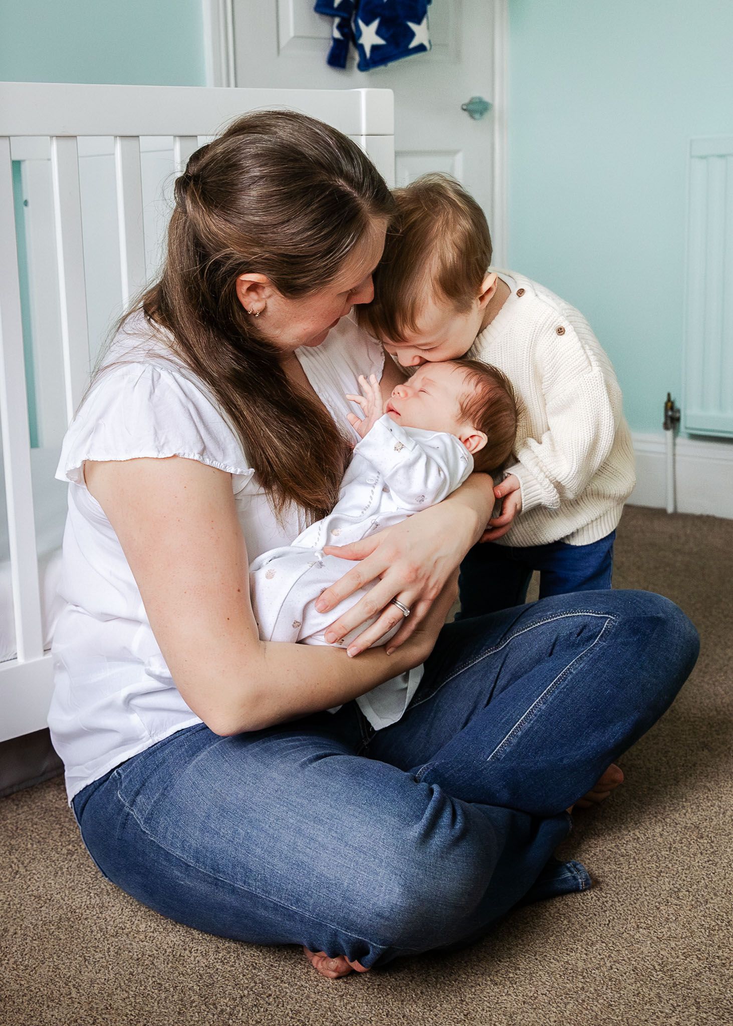 A toddler kisses his newborn brother on the head as the baby sleeps in his mum's arms. Taken during a relaxed photoshoot at home in Bristol.