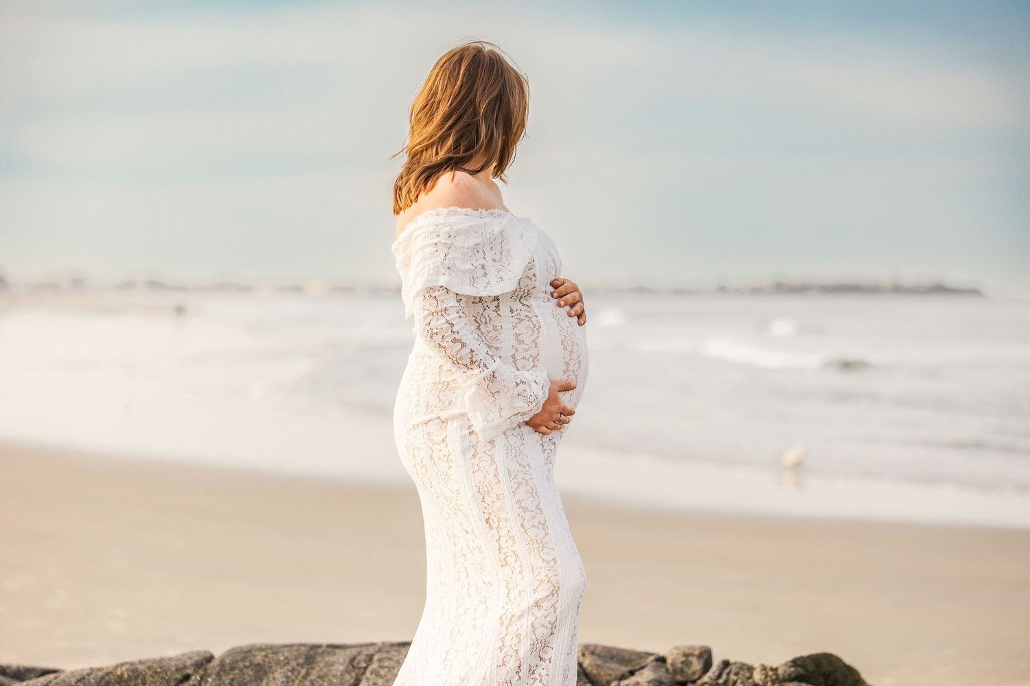 Pregnant woman in white lace dress holding baby bump on beach, maternity photo shoot by the ocean.