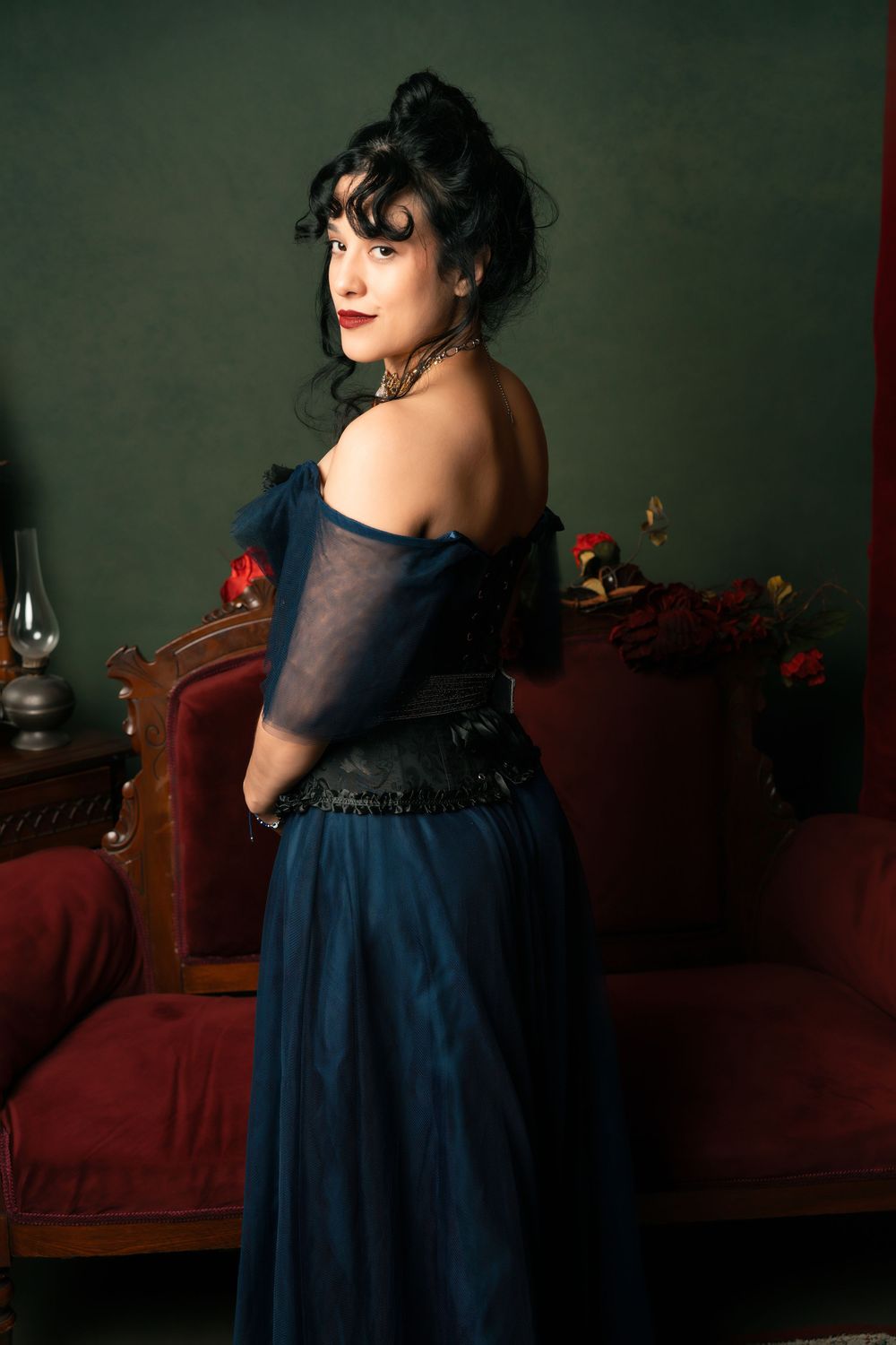 Woman in elegant navy blue off-shoulder gown poses in vintage Victorian-style room with red velvet furniture.
