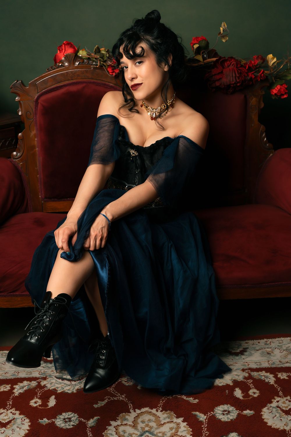 Woman in dark blue off-shoulder gown and black boots seated on red velvet antique sofa with roses.