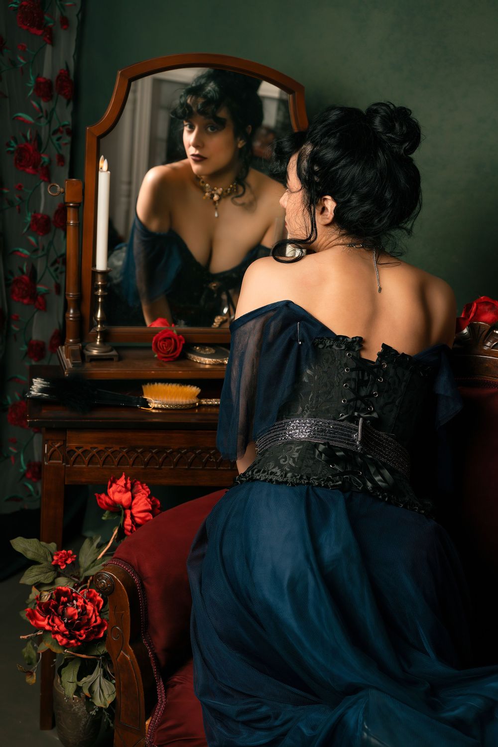 Woman in black corset and blue gown sits before vintage mirror with candle and roses in gothic Victorian setting.