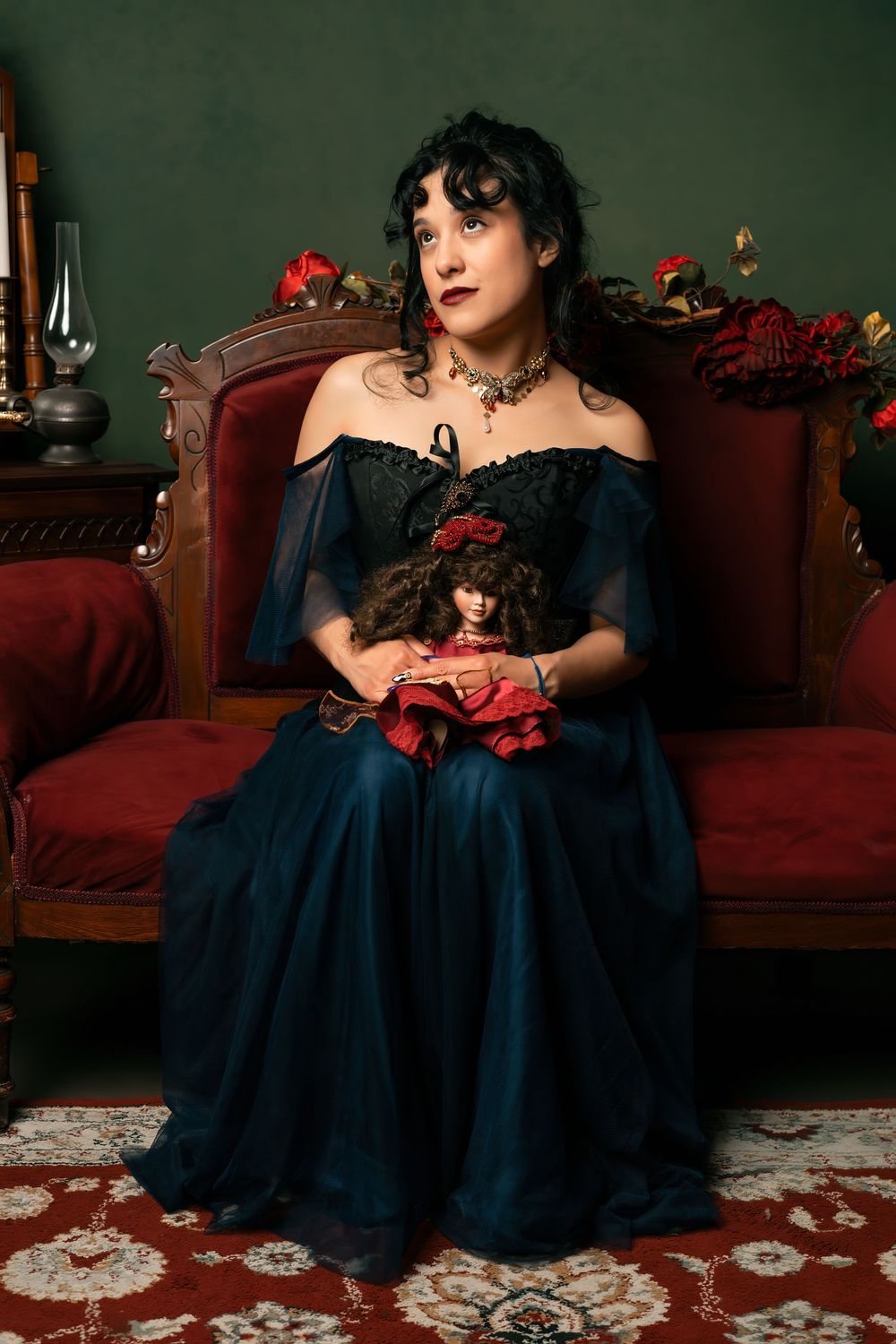 Woman in dark blue Victorian gown holds doll on red velvet sofa with roses and vintage decor.