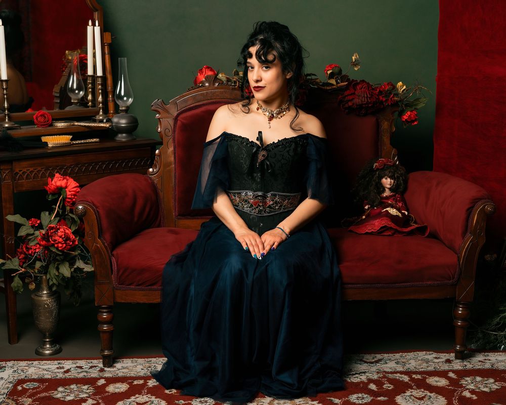 Woman in dark gothic gown seated on red velvet chair surrounded by roses and candles in moody Victorian setting.