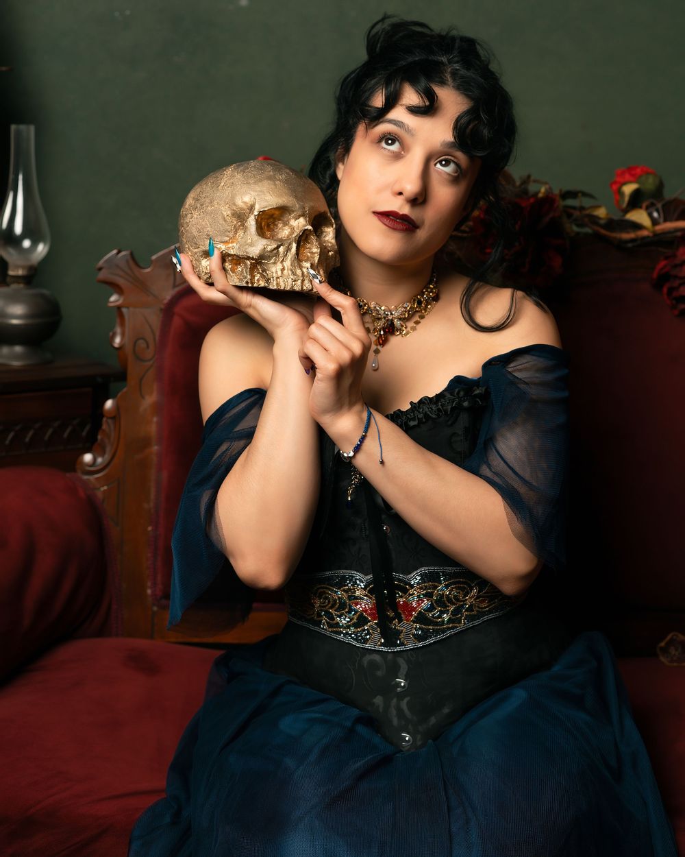 A gothic woman in a blue corset gown holds a skull while seated on a red velvet chair in a dark, dramatic setting.