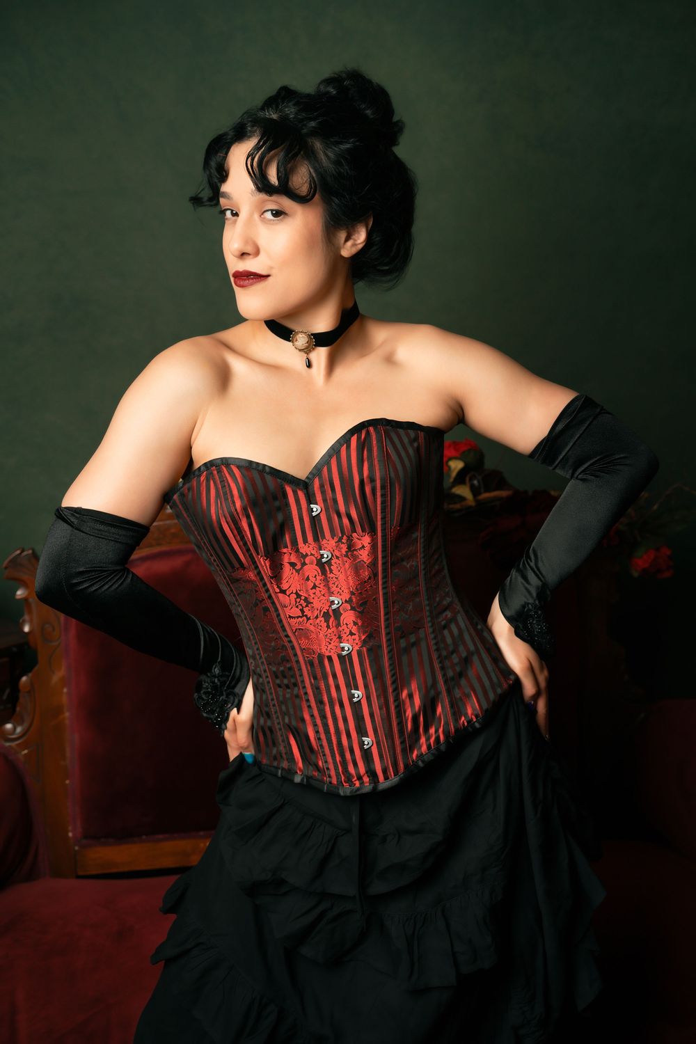 Woman in red and black striped corset with black gloves and skirt posing elegantly in a vintage-style setting.