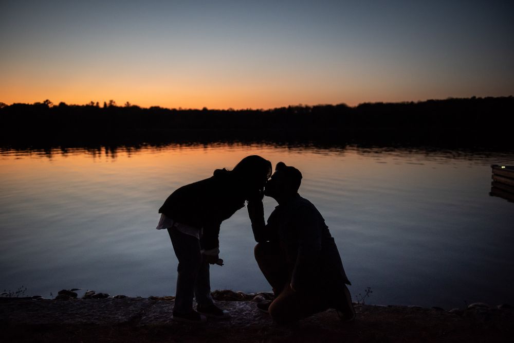 Silhouette of a man proposing to a woman by a lake at sunset.