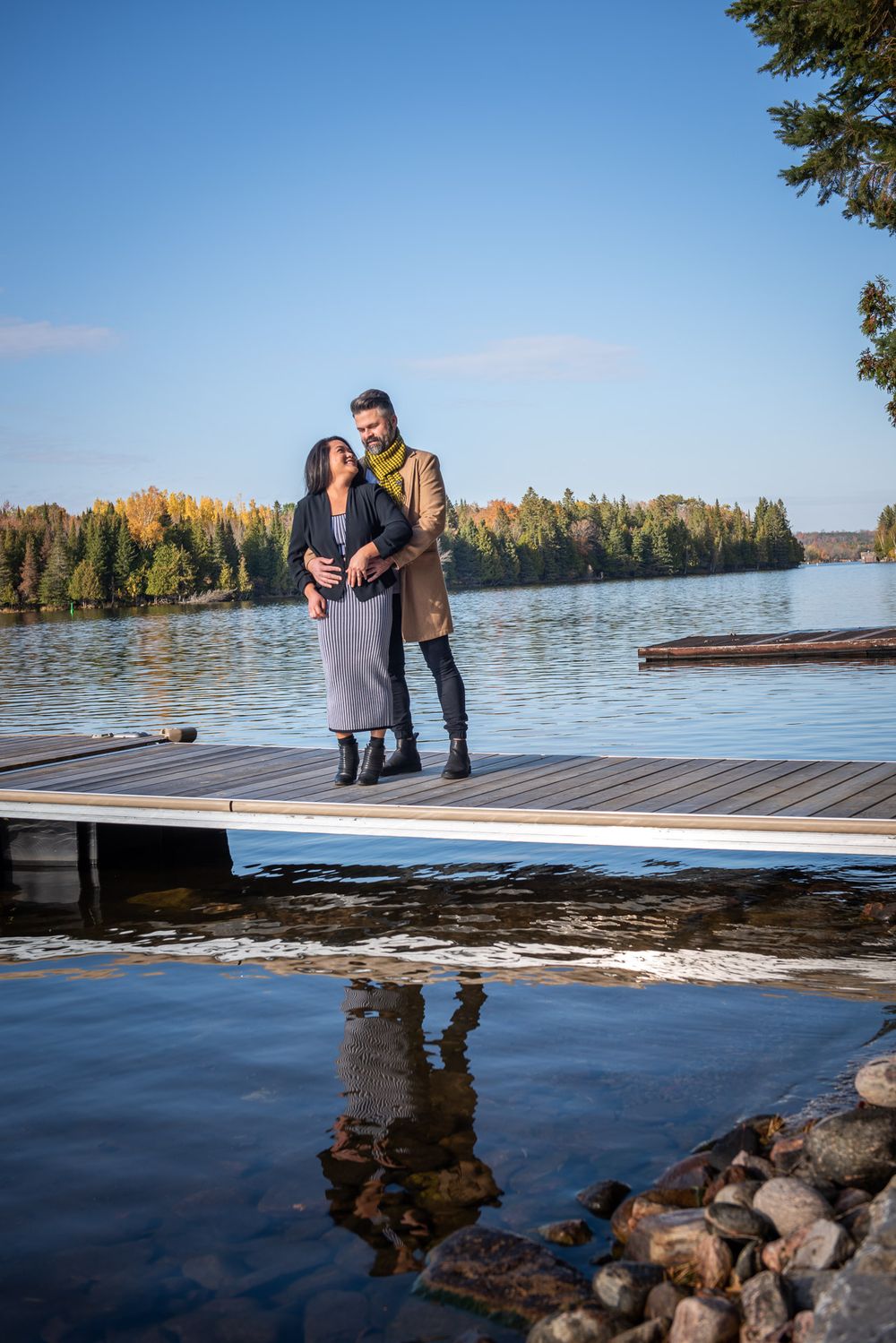 Couple embraces on a lakeside dock with stunning autumn foliage and blue sky reflecting on the water.
