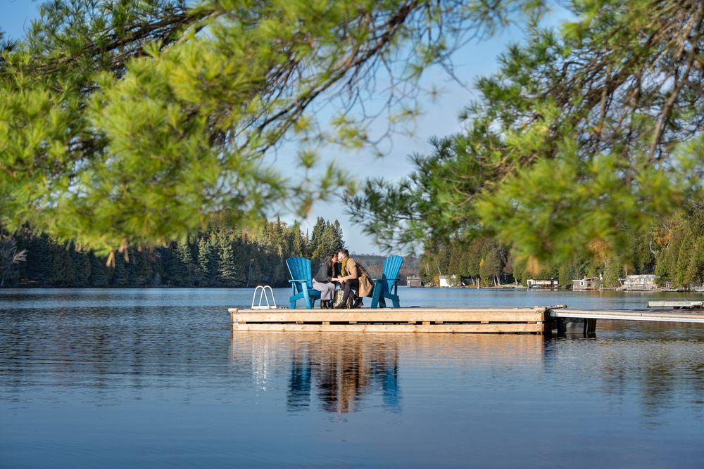 Couple sits in blue Adirondack chairs on a serene lake dock framed by lush green pine trees.