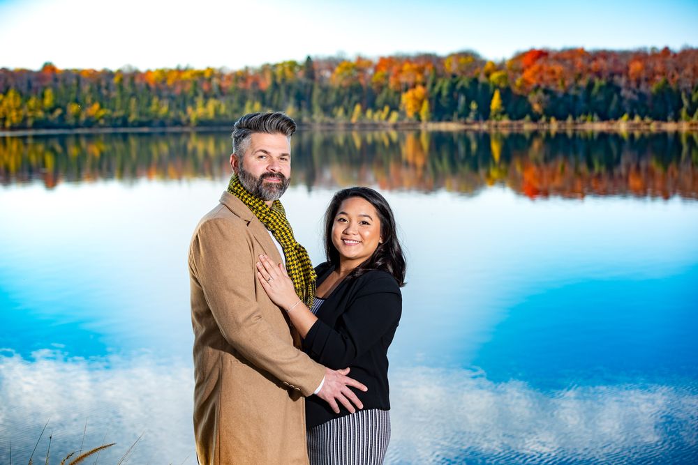 Couple smiles together by a lake with vibrant fall foliage perfectly reflected in the calm water.