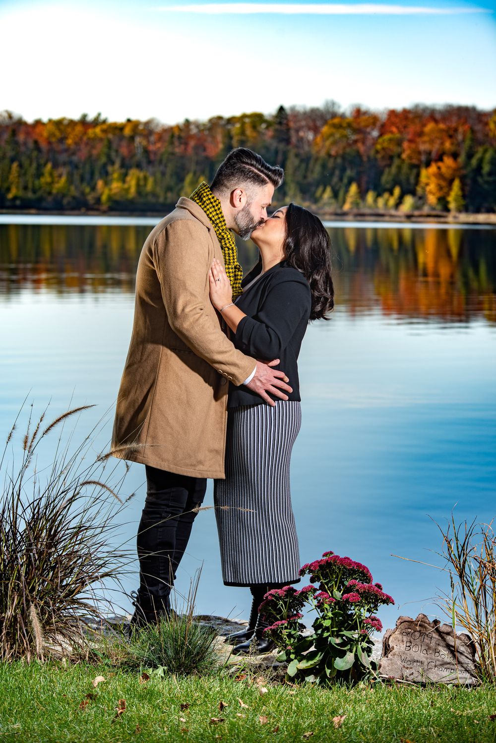 Couple shares a kiss beside a tranquil lake with colorful autumn trees and a red flower nearby.