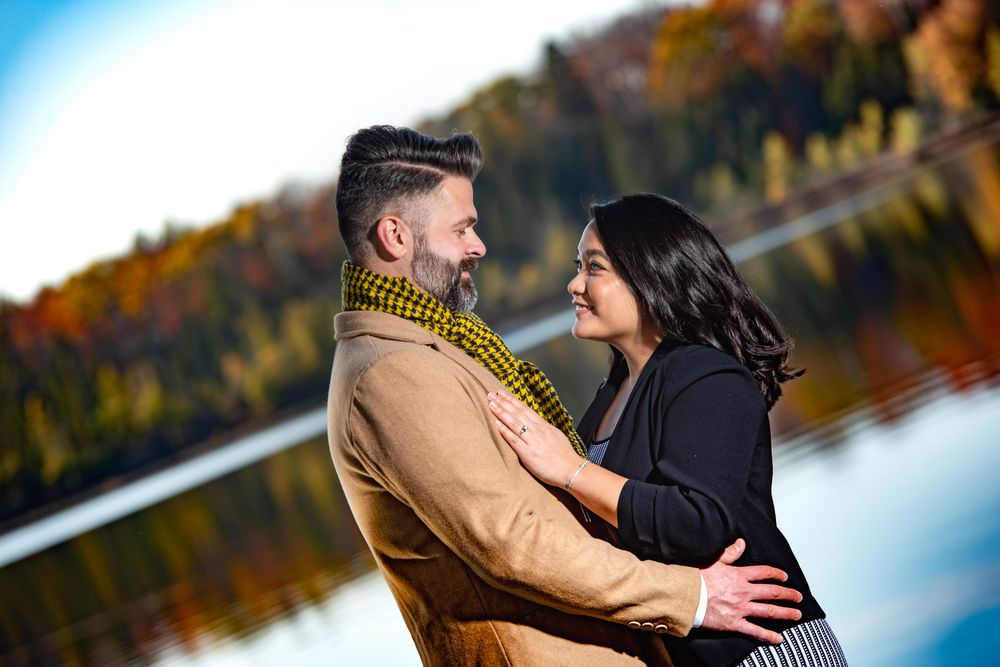 Couple gazes lovingly at each other by a lakeside with brilliant fall foliage in the background.