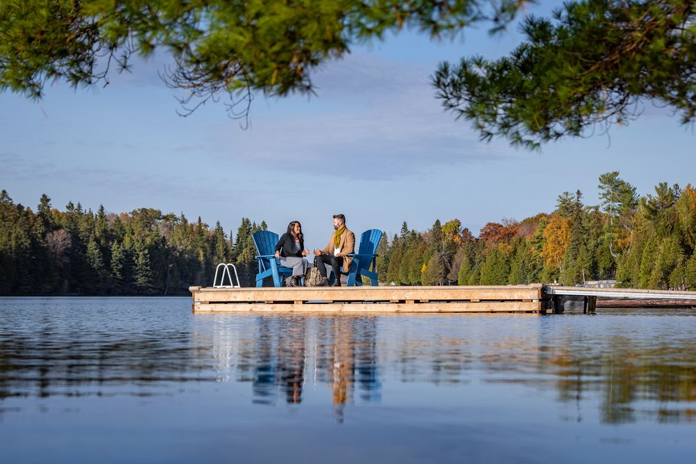 Couple relaxes on a wooden dock surrounded by calm lake waters and colorful autumn forest reflections.