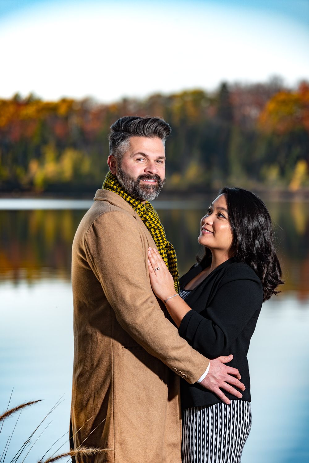 Bearded man and woman share a tender embrace by a lake with colorful autumn trees behind them.