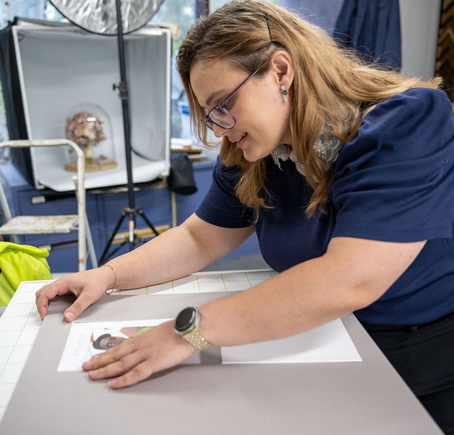 Woman in blue shirt examines document on white table in professional studio setting with photography equipment.