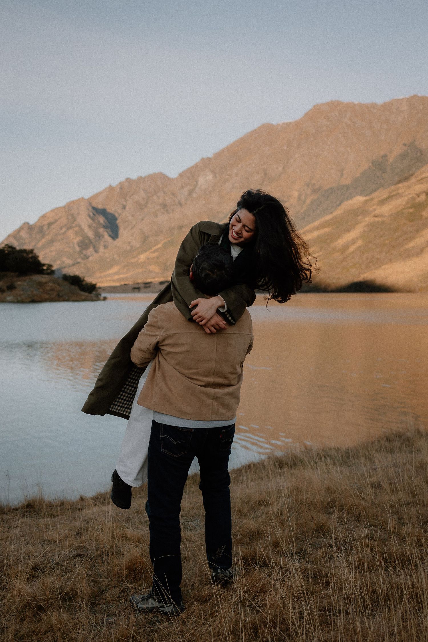Couple embracing and laughing in a mountain field at sunset, woman in olive coat, man in tan jacket.