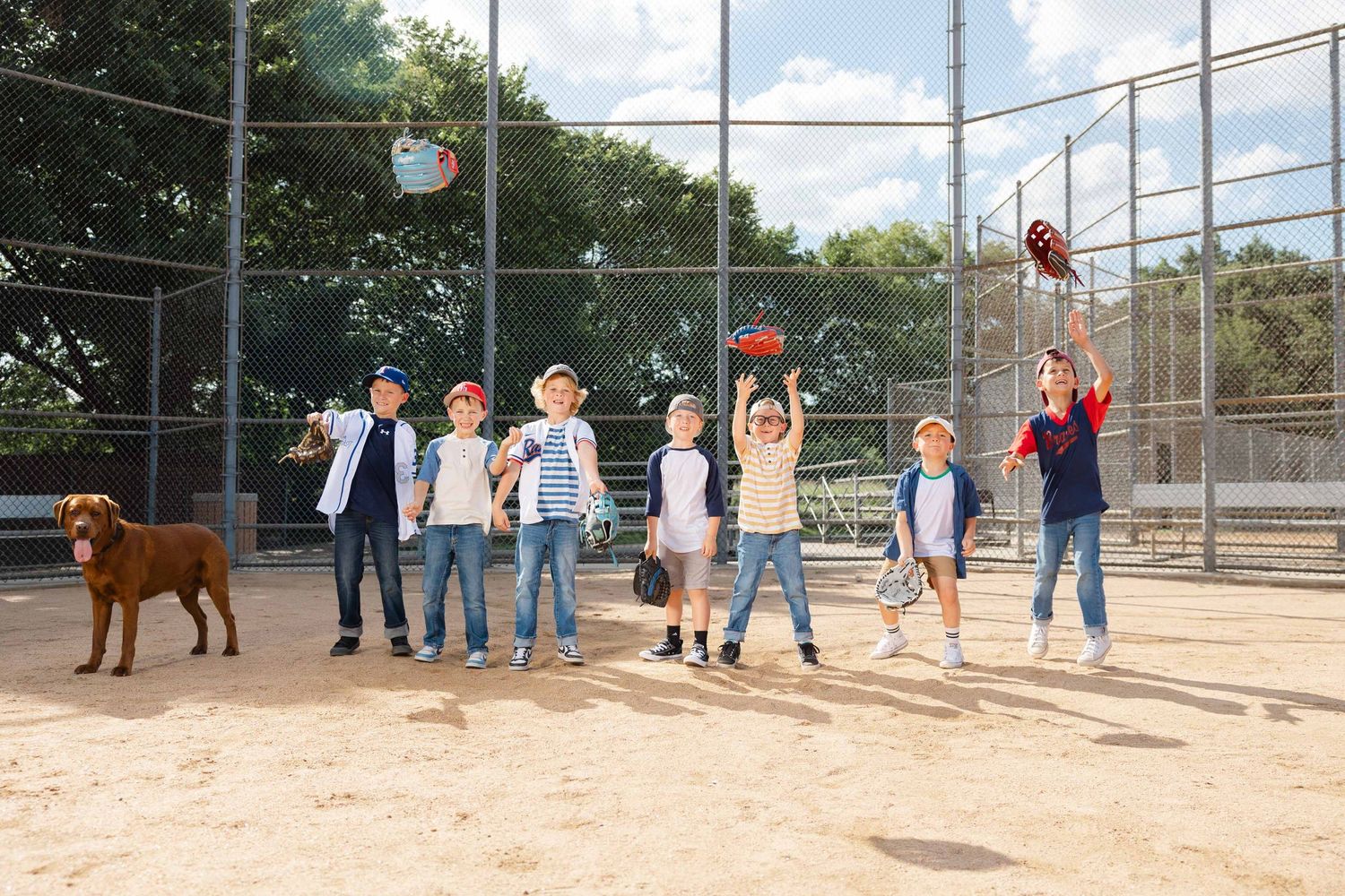 Kids playing baseball on a sandy field with a dog, tossing balls and having fun behind a chain-link fence.