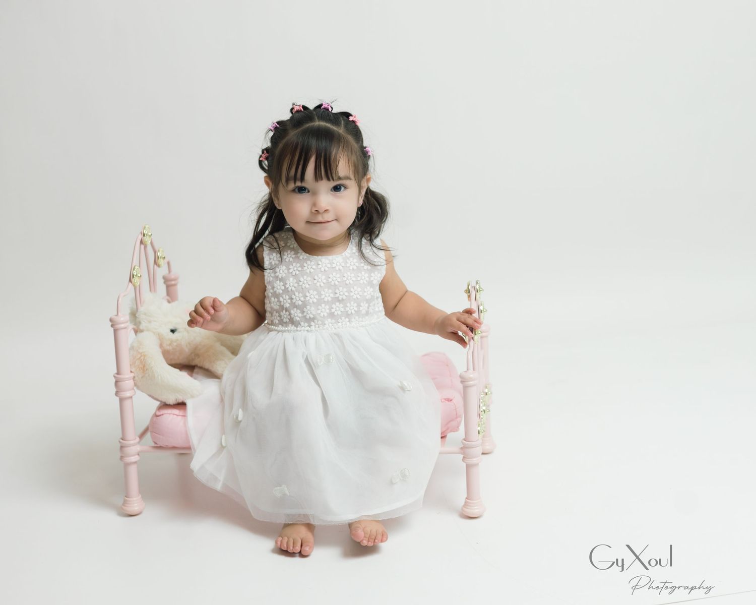 Toddler girl in white dress sitting on pink miniature bed for professional photo session.