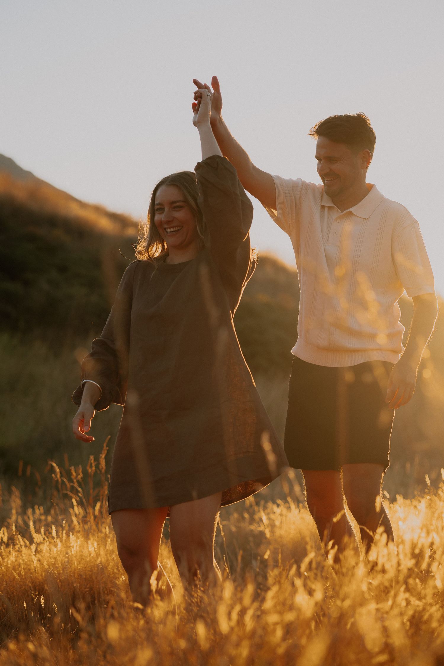 Happy couple embracing in golden field at sunset with mountain backdrop, woman in brown dress laughing joyfully.