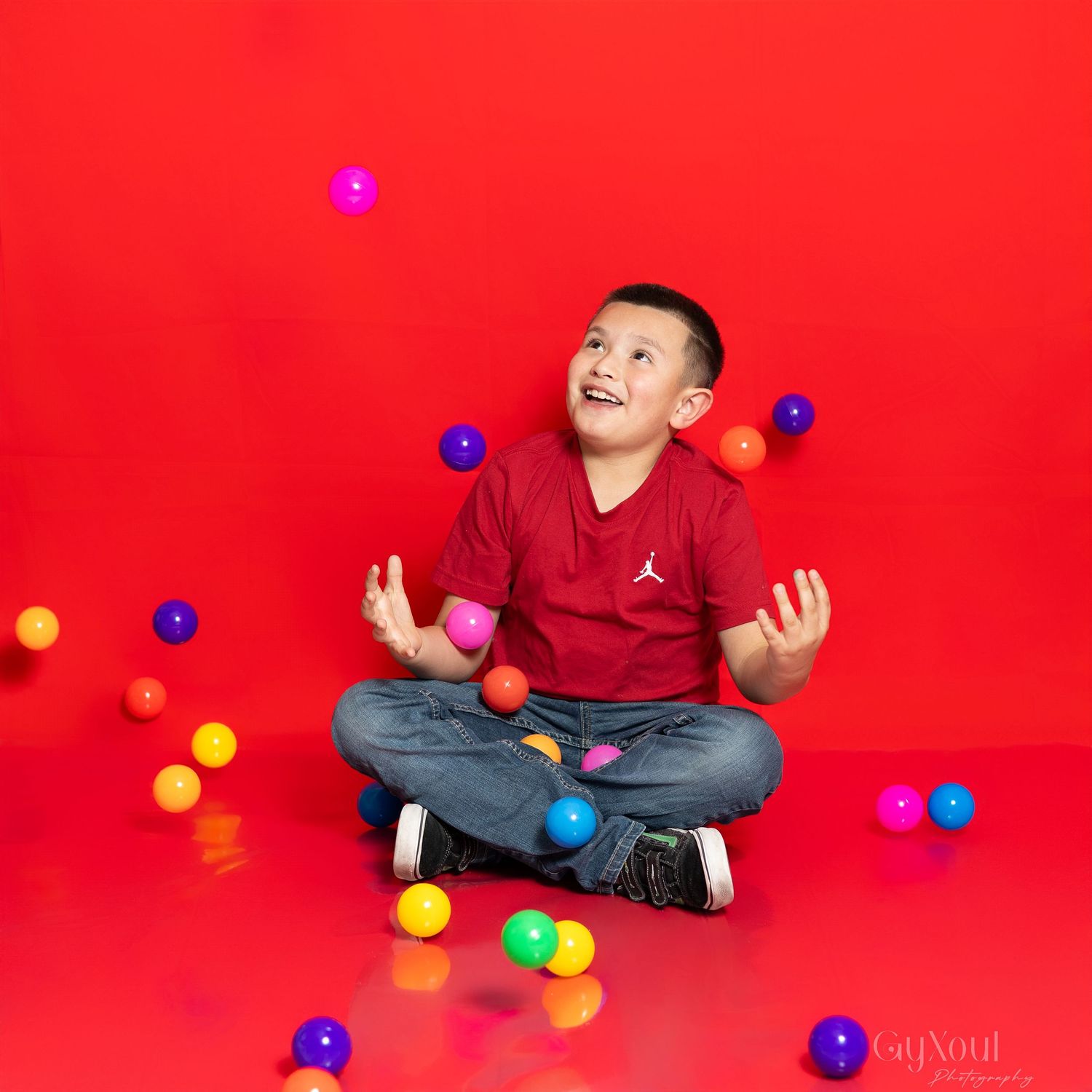 A young boy in a red Jordan shirt sits cross-legged on a red floor, joyfully tossing colorful balls in the air.