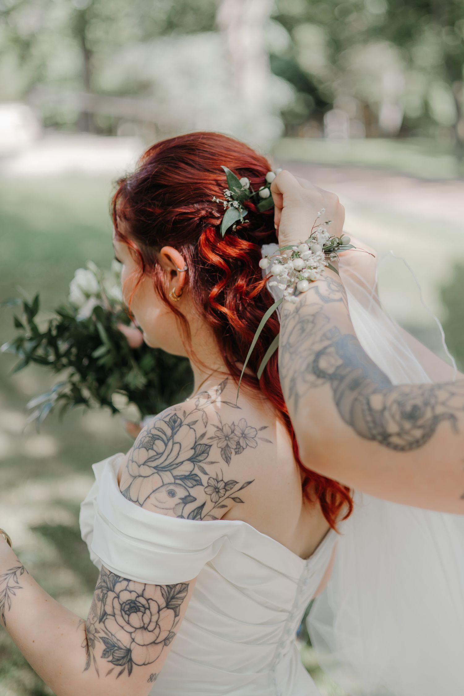 Tattooed bride in white off-shoulder gown with red updo adorned with flowers holds bouquet outdoors.