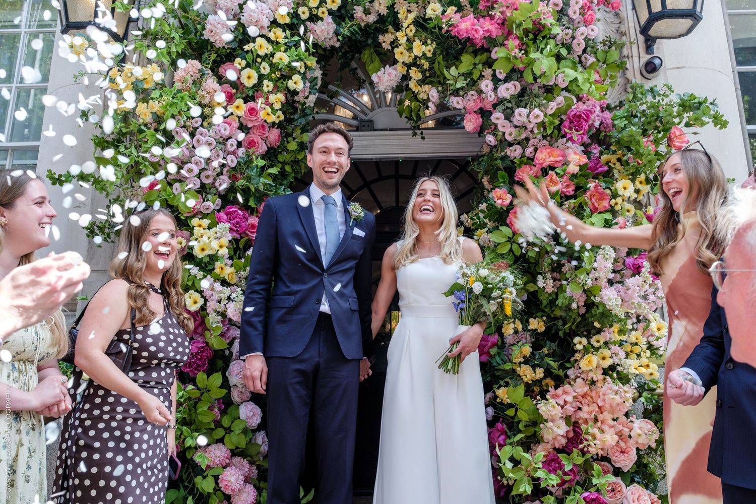 Bride and groom exit under a stunning floral arch surrounded by guests throwing confetti at an outdoor wedding celebration.