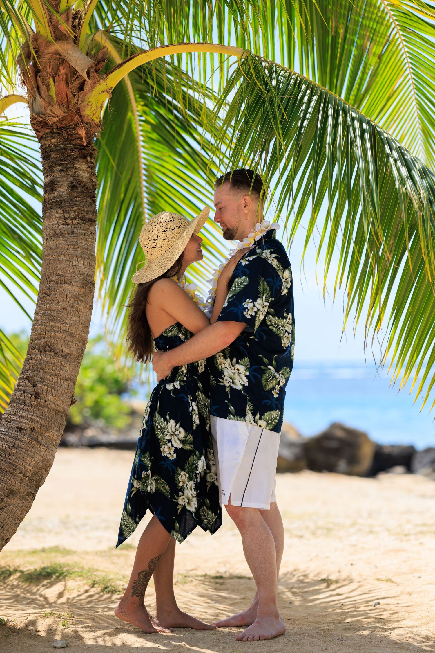 Romantic couple in matching Hawaiian floral outfits embracing under a palm tree on a tropical beach at Waialae beach park