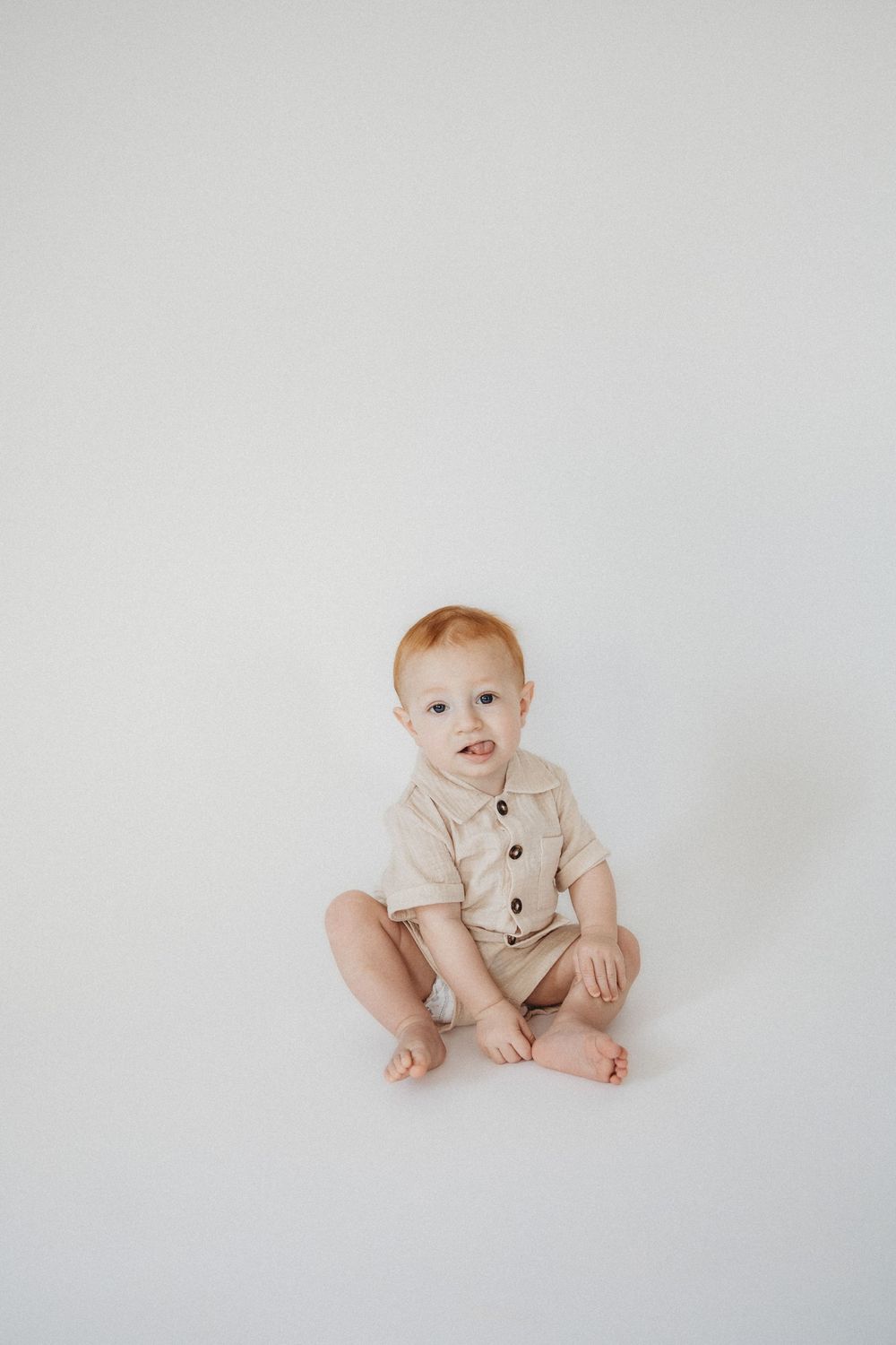 A cute baby with red hair sits on a white background wearing a beige button-up romper, looking at the camera.