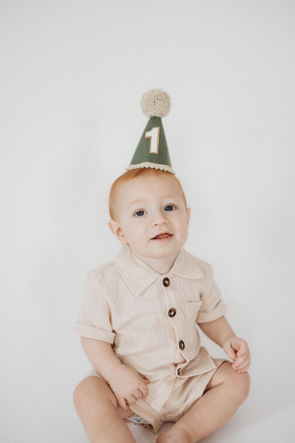 Smiling baby wearing a green felt number 1 birthday hat and beige romper sitting against a white background.
