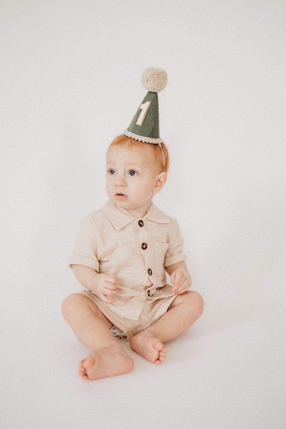 Cute baby wearing a green number 1 birthday hat and beige romper sitting on white background.